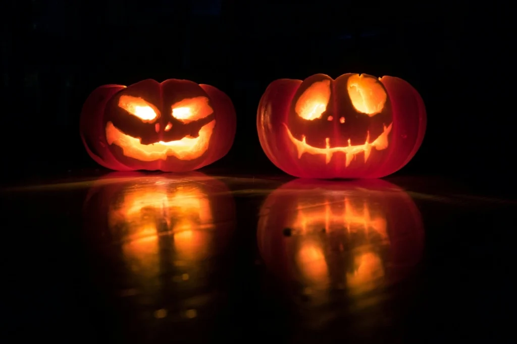 Mustalla taustalla kaksi kaiverrettua, sisältä valaistua Halloween-kurpitsaa.
