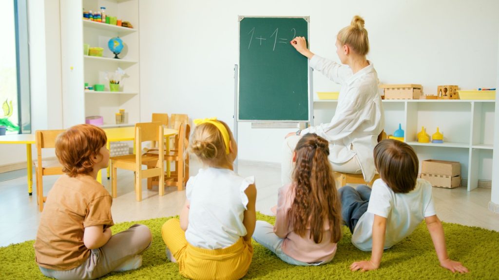 Dzieci siedzą w klasie podczas zajęć z nauczycielką przy tablicy, prezent na koniec roku szkolnego.