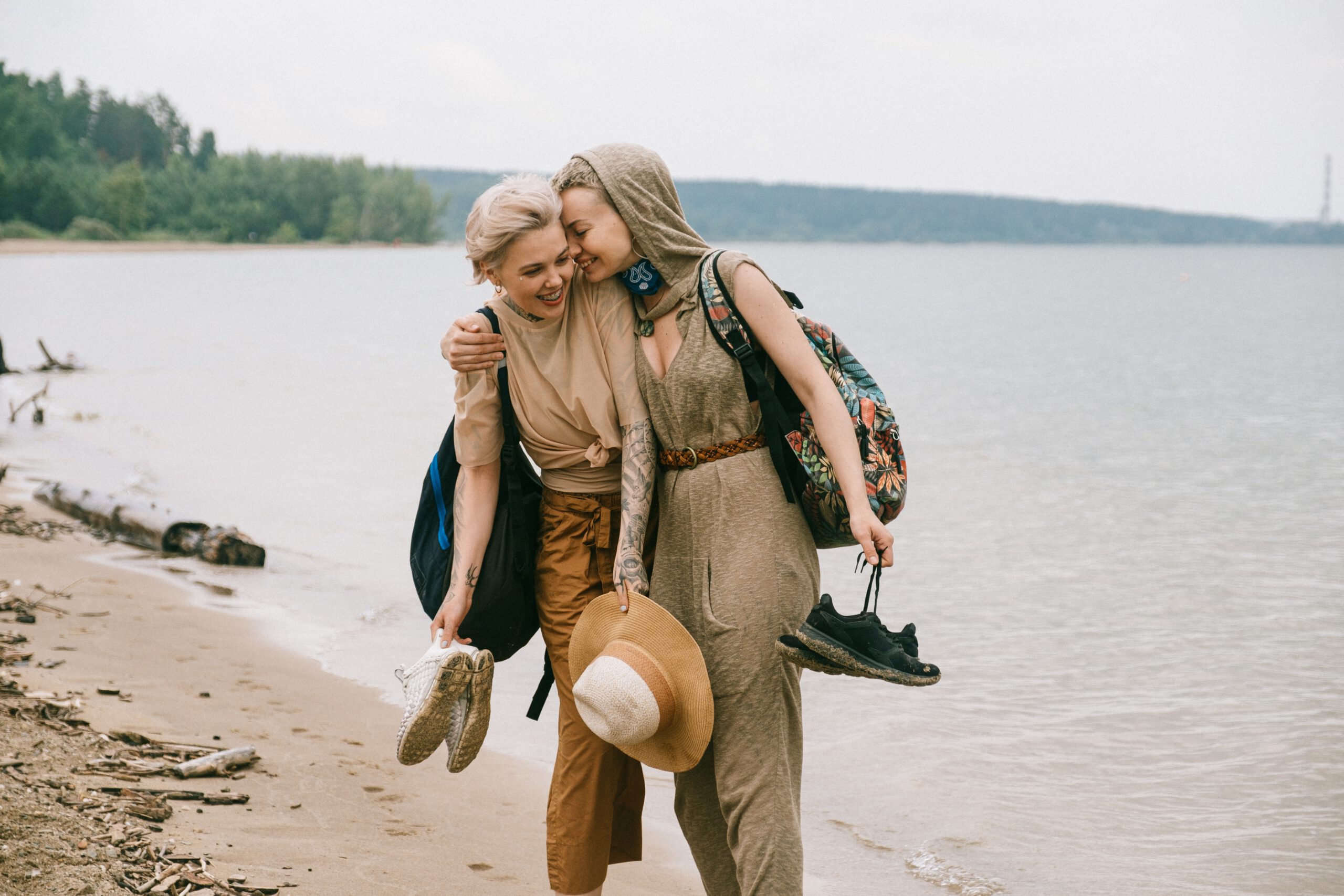 Kobiety idą, przytulając się. Trzymają w dłoniach buty. Idą po plaży. W tle jezioro i wzgórza.