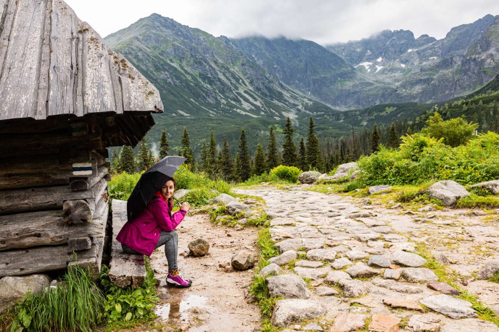 Mniej uczęszczane szlaki w Tatrach - kobieta w różowej kurtce przeciwdeszczowej siedzi pod parasolką, pod dachem bacówki, przy szlaku w górach, w tle Tatry za chmurami