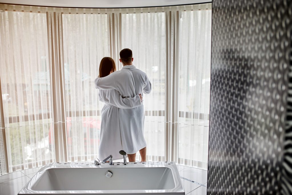 Hotel SPA dla Dwojga - para w szlafrokach, odwrócona plecami, stoi w objęciach, przy oknie, patrząc na zewnątrz.