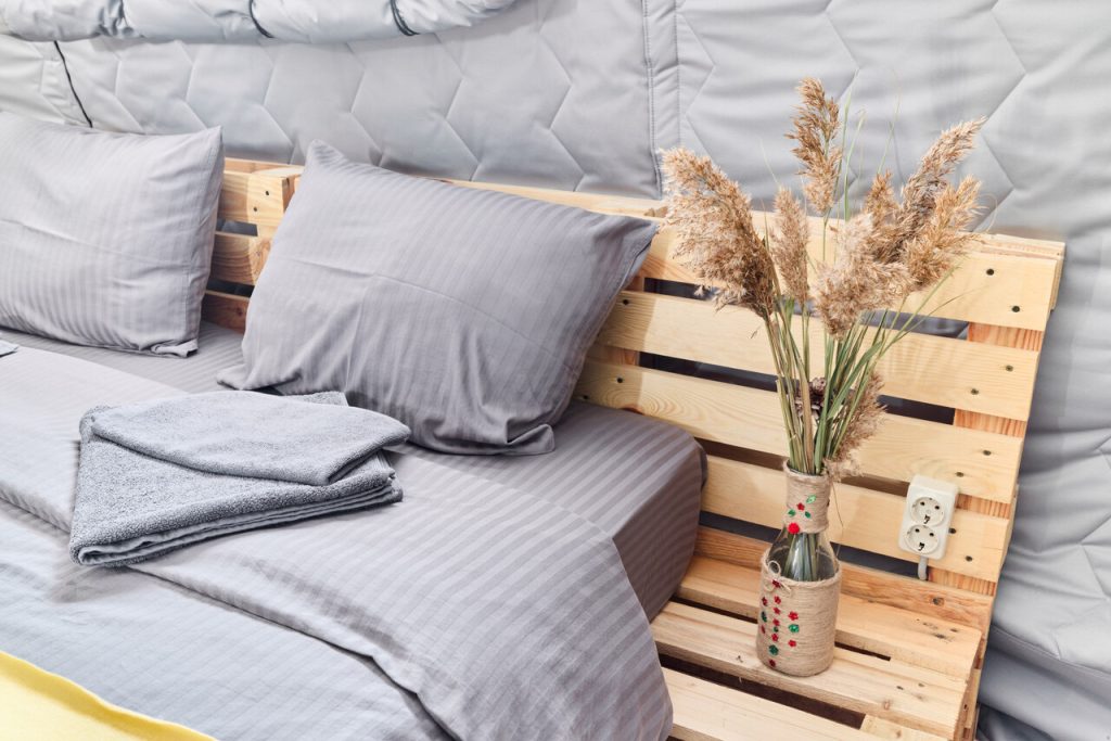 Wnętrze glampingu w Polsce – wygodne łóżko z szarą pościelą i drewnianym wezgłowiem z palet, obok dekoracja z suszonych traw w wazonie.
