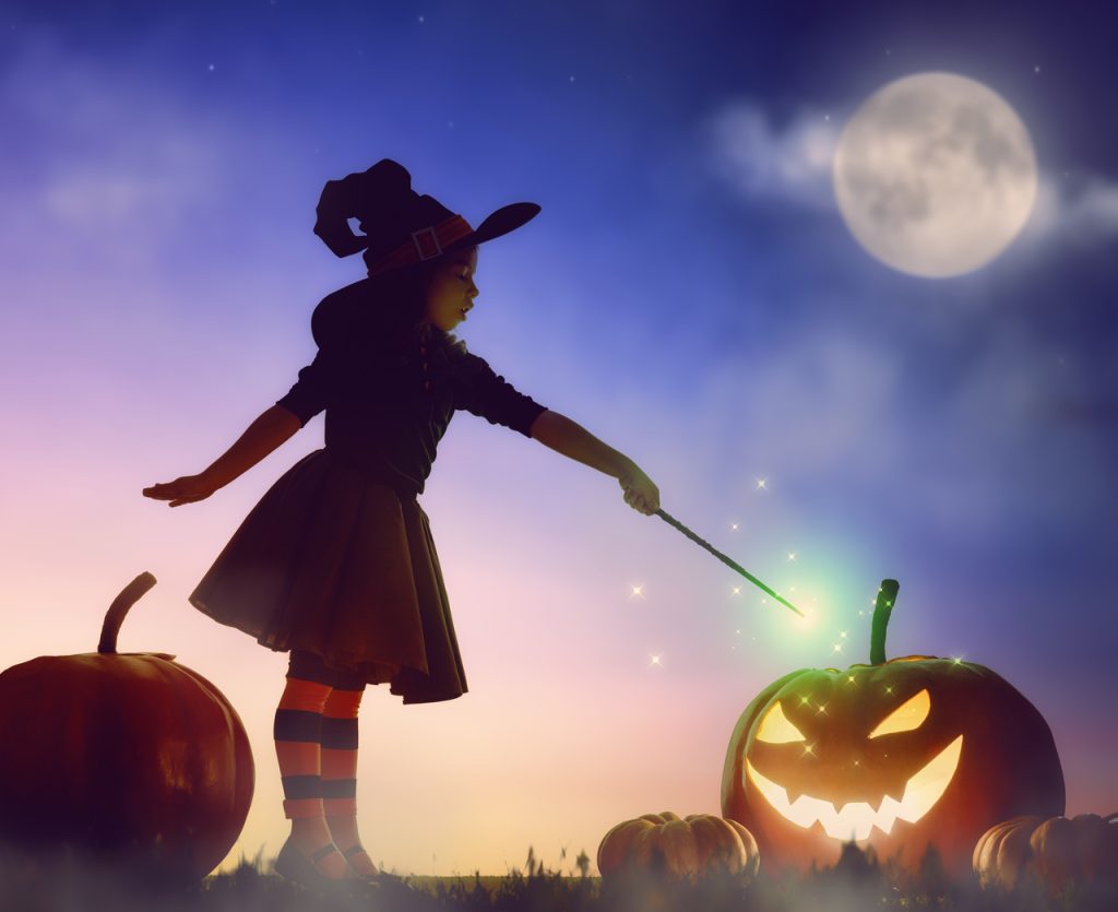 Dziewczynka przebrana za czarownicę z magiczną różdżką oświetla dynię Jack O’Lantern w blasku księżyca — nastrojowa scena symbolizująca magię Halloween obchodzonego 31 października.