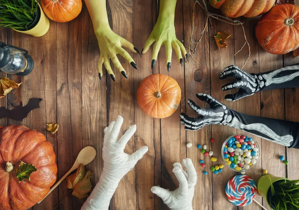 Stół na Halloween z dyniami i cukierkami, po które sięgają ręce w kostiumach potworów i mumii.