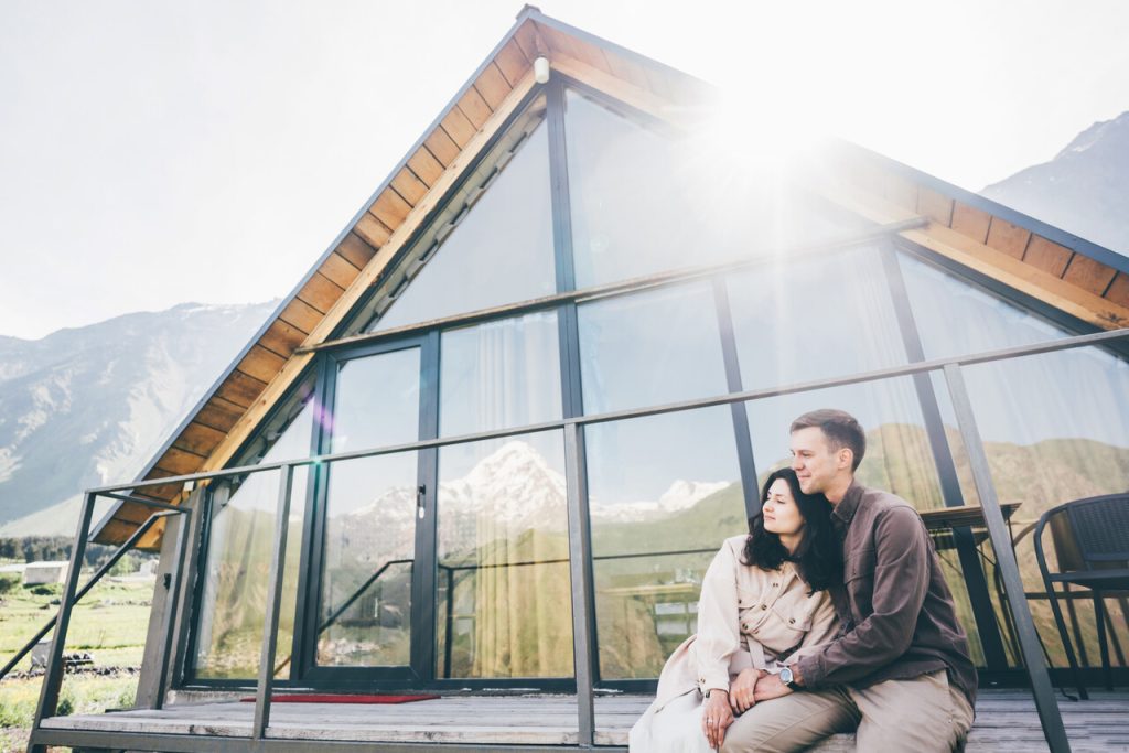 Para odpoczywająca przed nowoczesnym domkiem w górach z panoramicznym widokiem na ośnieżone szczyty. Idealne miejsce na romantyczne wakacje zimą lub jesienią w naturze.