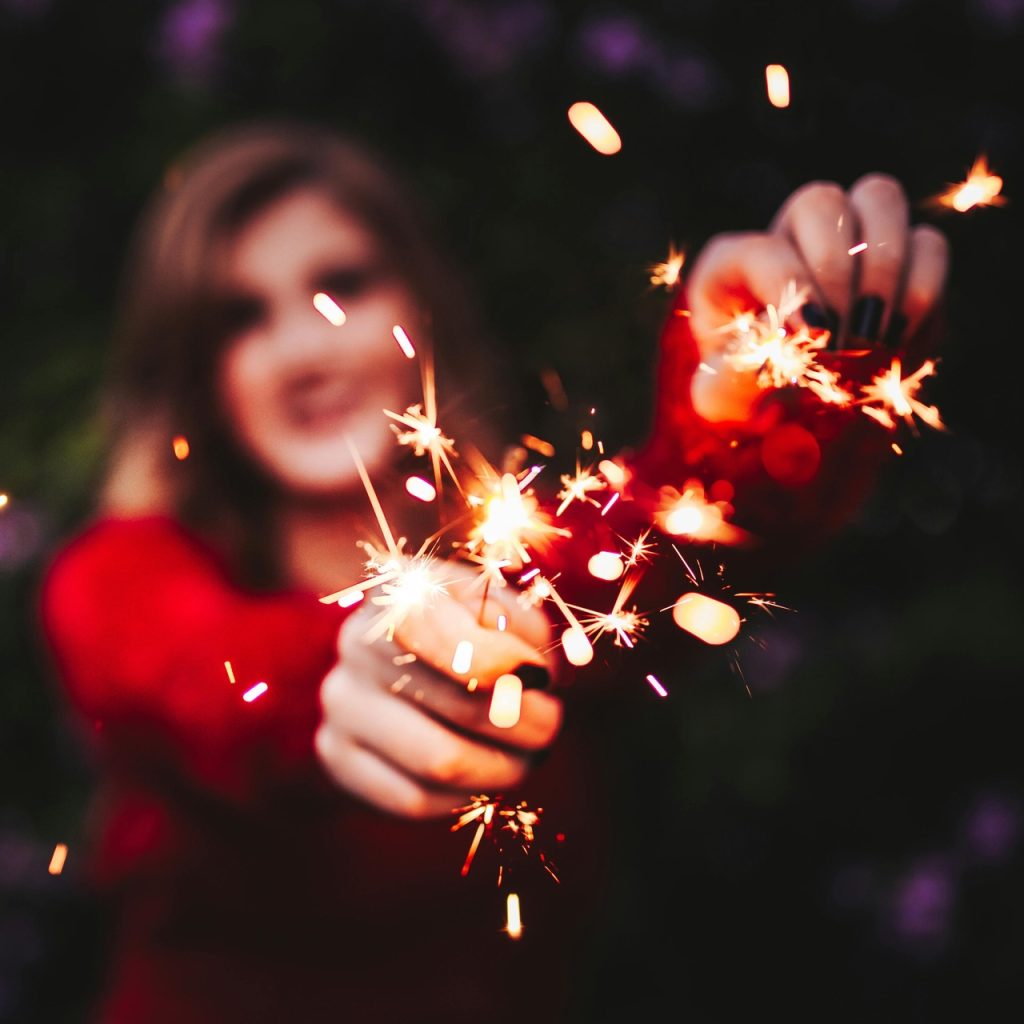 Kobieta w czerwonym swetrze trzyma w dłoniach zapalone zimne ognie, tworząc ciepły, iskrzący kadr idealny do tematu kiedy są Katarzynki i listopadowych zwyczajów wróżebnych.