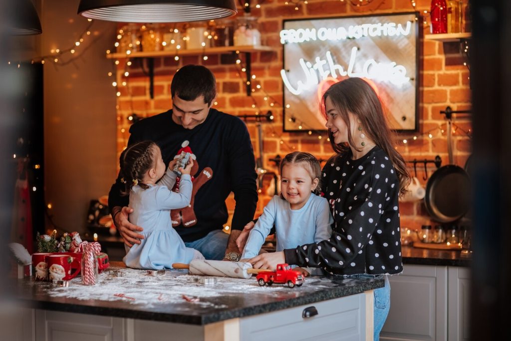Rodzina wspólnie przygotowuje świąteczne wypieki w kuchni ozdobionej lampkami i dekoracjami, a dzieci stoją przy blacie obsypanym mąką.