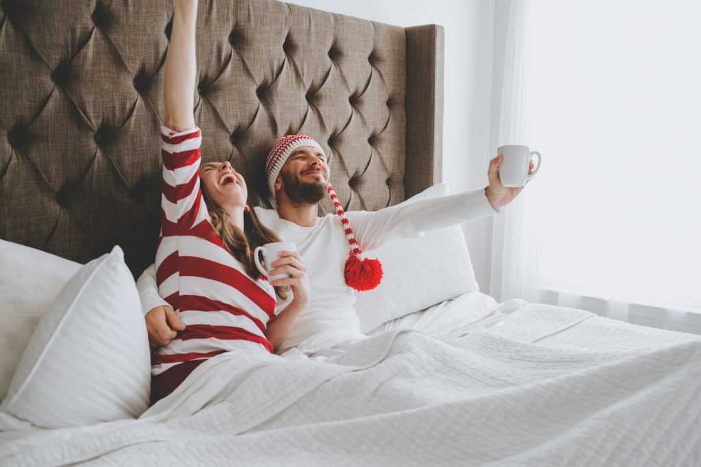 Para siedzi w łóżku pod białą kołdrą, trzymając kubki i ciesząc się świątecznym porankiem; jedna z osób ma na głowie czapkę z pomponem.