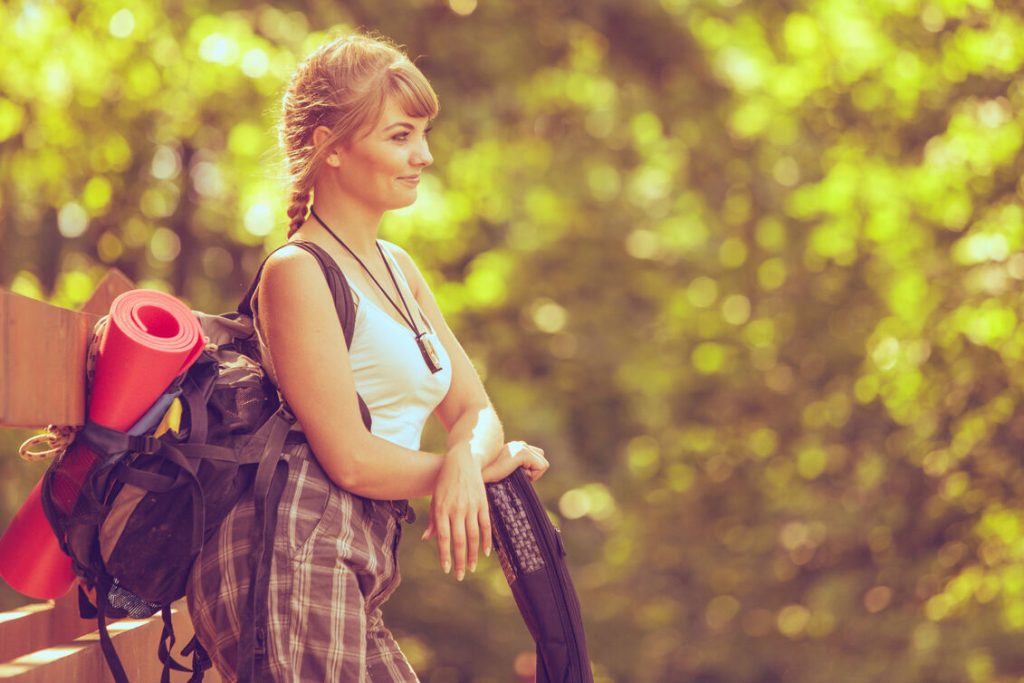 Kobieta z dużym plecakiem i karimatą siedzi na leśnej ławce podczas wędrówki — kadr oddaje klimat prezentu dla podróżnika i miłośniczki outdooru.