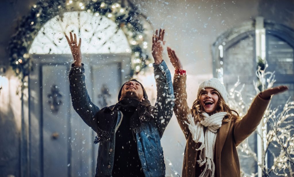 Dwie osoby ubrane w zimowe kurtki i czapki stoją na udekorowanej światłami ulicy, unosząc ręce i ciesząc się spadającym śniegiem.